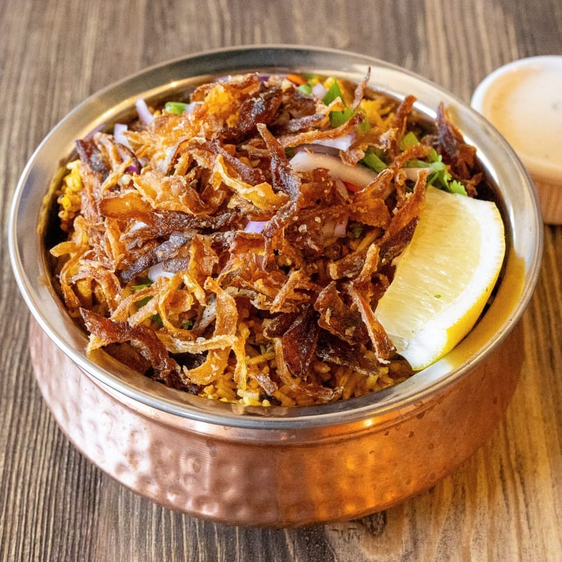 Biryani in a copper bowl, topped with crispy fried onions, fresh herbs, and a lemon wedge.