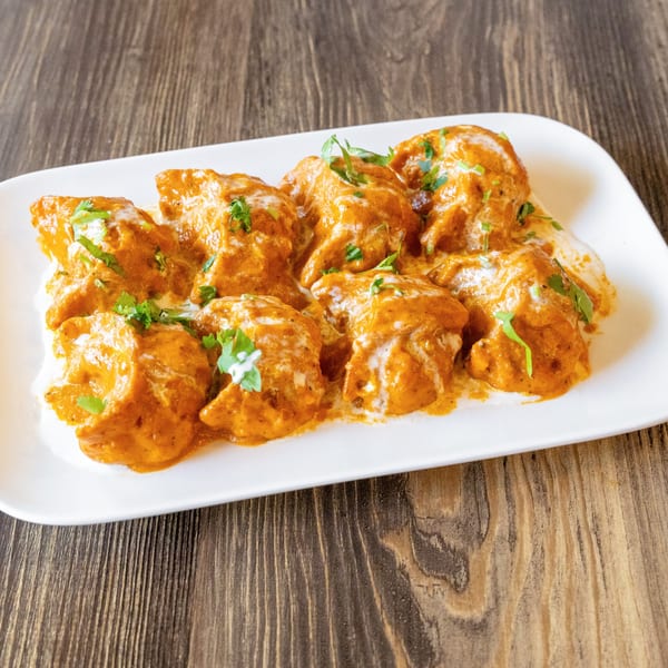 A white rectangular plate filled with momos in orange sauce, drizzled with white cream and green herbs.
