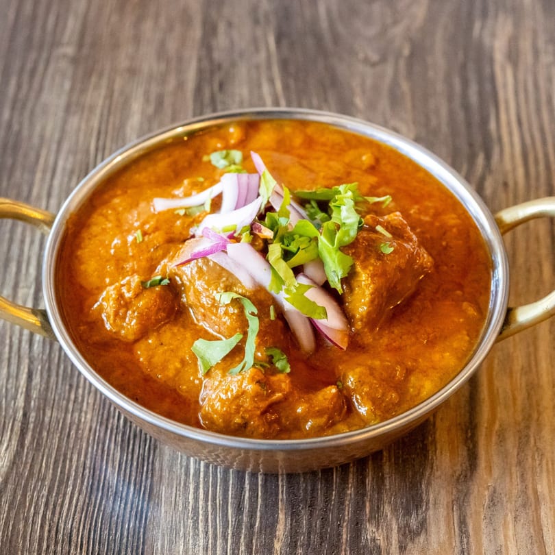 A copper bowl of chicken curry, garnished with red onions and cilantro, on a wooden table.
