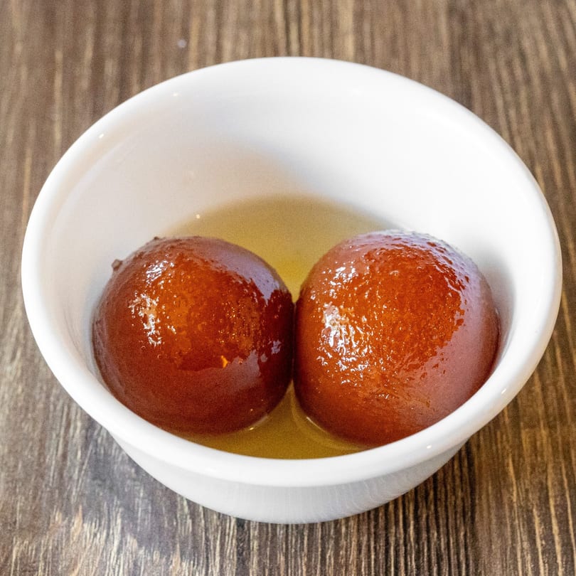 Two glistening gulab jamun in a white bowl, submerged in golden syrup on a wooden table.