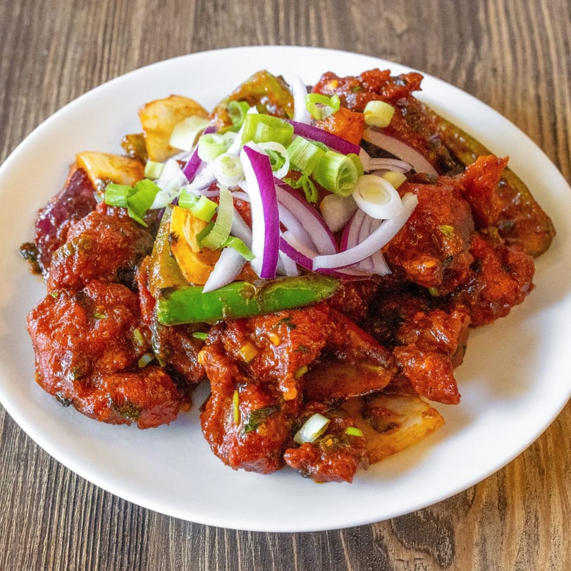 A white plate of chili chicken, garnished with sliced red onions, green onions, and green chilies.