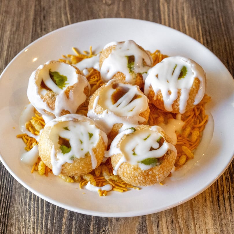 Six pani puri shells filled with green chutney and drizzled with white yogurt sauce on a bed of sev.