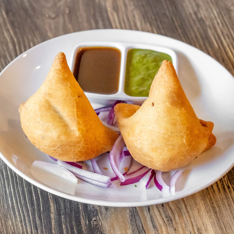 Two golden-brown samosas on a white plate with red onion, green and brown dipping sauces.