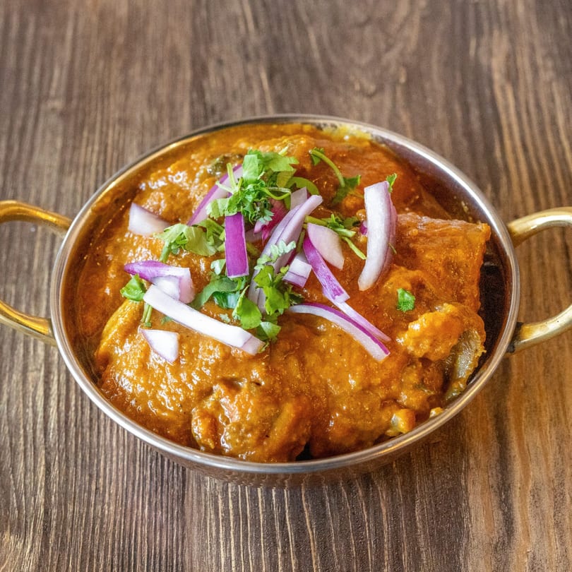 Chicken curry in a brass bowl, garnished with red onions and cilantro, on a wooden table.
