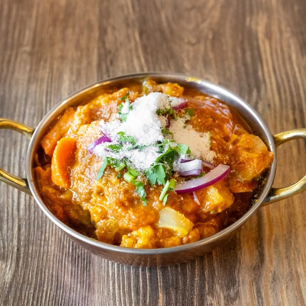 A metal bowl of Indian curry with vegetables, topped with shredded coconut, red onions, and cilantro.